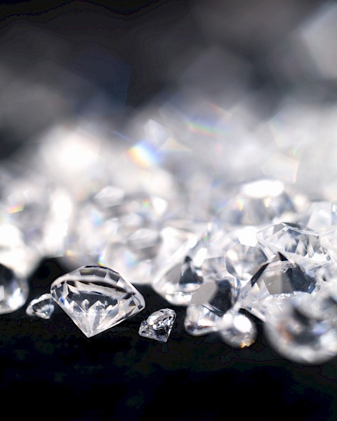 The image displays a collection of sparkling diamonds scattered on a dark surface, with some out of focus in the background.