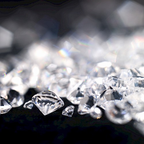 The image displays a collection of sparkling diamonds scattered on a dark surface, with some out of focus in the background.