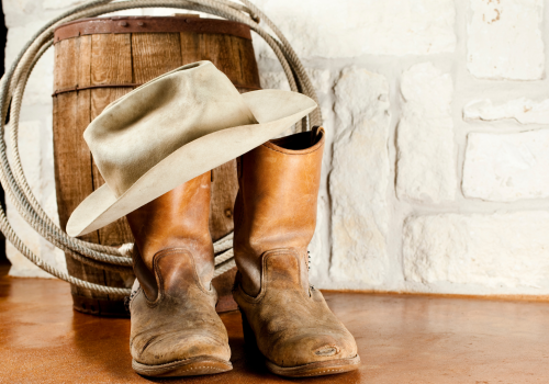 The image shows a pair of worn cowboy boots, a cowboy hat resting on top, a coiled rope hung on a barrel, and a white brick wall in the background.