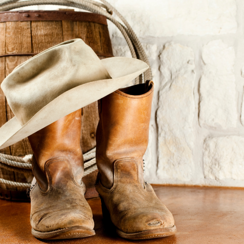 The image shows a pair of worn cowboy boots, a cowboy hat resting on top, a coiled rope hung on a barrel, and a white brick wall in the background.