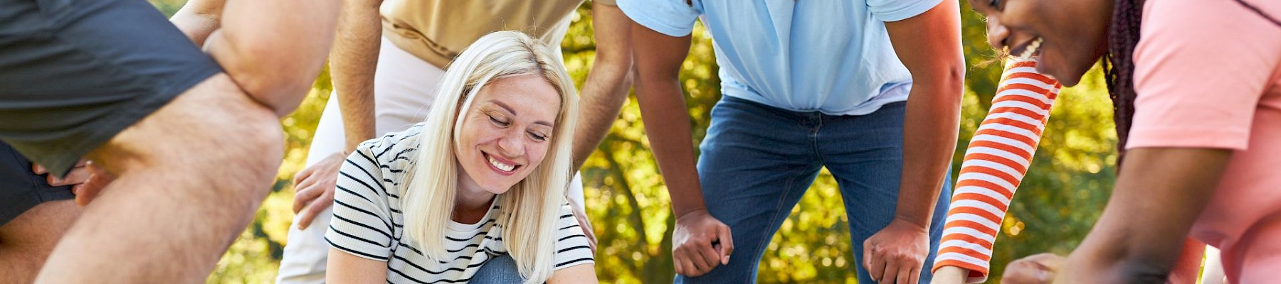 A group of people are gathered outdoors, leaning over and pointing at something on a white cloth on the grass, appearing focused and engaged.