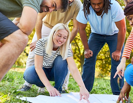 A group of people are gathered outdoors, leaning over and pointing at something on a white cloth on the grass, appearing focused and engaged.