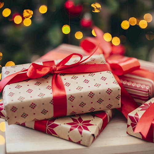 The image shows wrapped gifts with red ribbons, placed on a table with a decorated Christmas tree and twinkling lights in the background.