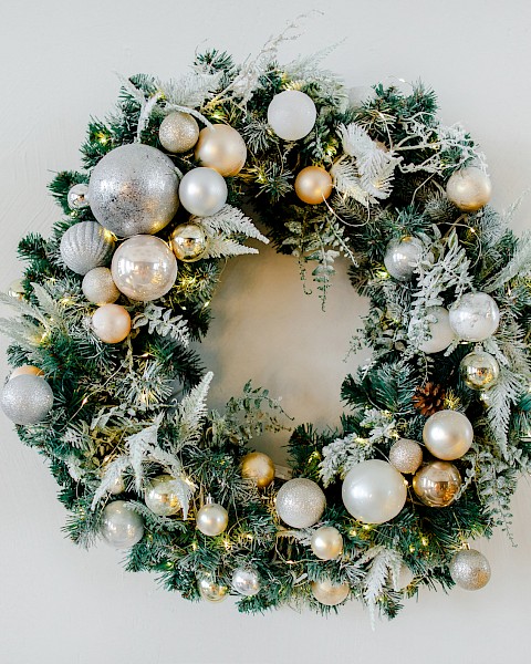 This is an image of a holiday wreath decorated with silver and gold ornaments, pine cones, and frosted greenery against a plain background.