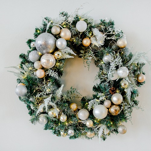 This is an image of a holiday wreath decorated with silver and gold ornaments, pine cones, and frosted greenery against a plain background.