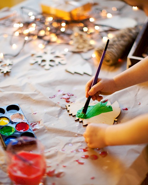 A child is painting a wooden ornament green using watercolors, with various painting supplies and wooden cutouts spread on the table.