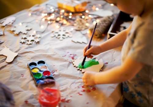 A child is painting a wooden ornament green using watercolors, with various painting supplies and wooden cutouts spread on the table.