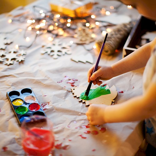 A child is painting a wooden ornament green using watercolors, with various painting supplies and wooden cutouts spread on the table.