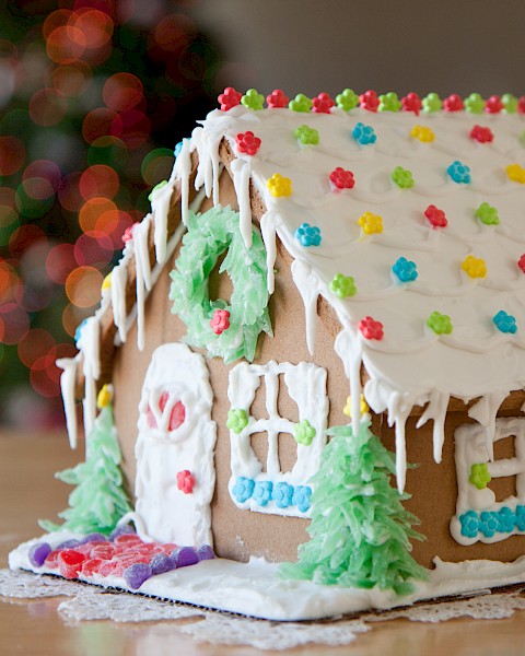 A decorated gingerbread house with colorful candies and white icing, situated on a table with a blurred festive background in view.