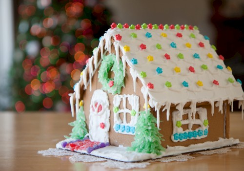 A decorated gingerbread house with colorful candies and white icing, situated on a table with a blurred festive background in view.