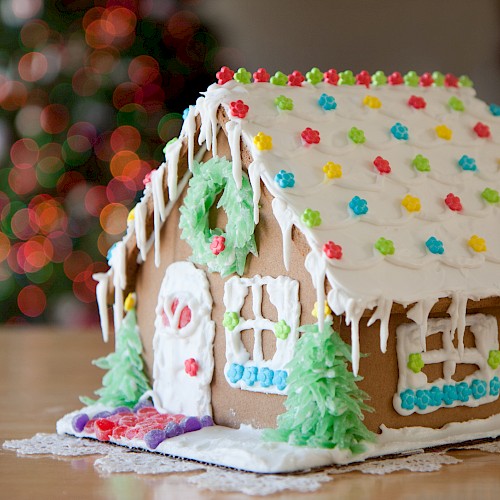 A decorated gingerbread house with colorful candies and white icing, situated on a table with a blurred festive background in view.