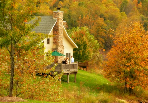 A cozy house with a stone chimney set in an autumn landscape; three people are standing on the wooden deck enjoying the view.