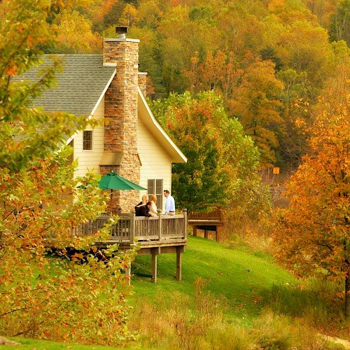 A cozy house with a stone chimney set in an autumn landscape; three people are standing on the wooden deck enjoying the view.