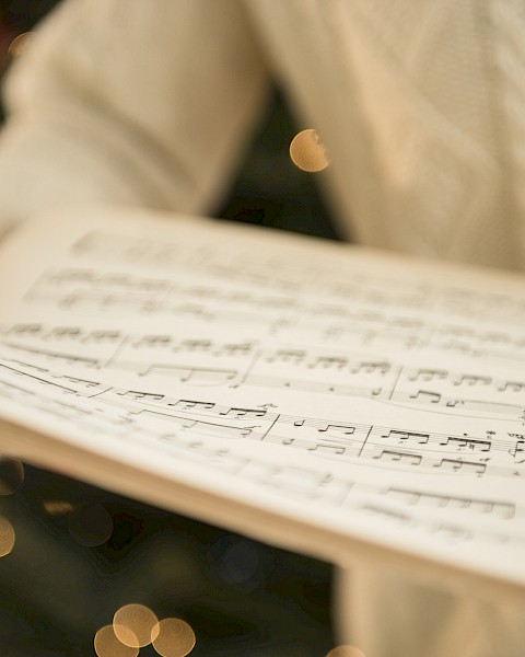 A person is holding an open book of sheet music, with out-of-focus festive lights in the background, possibly indicating a holiday season.