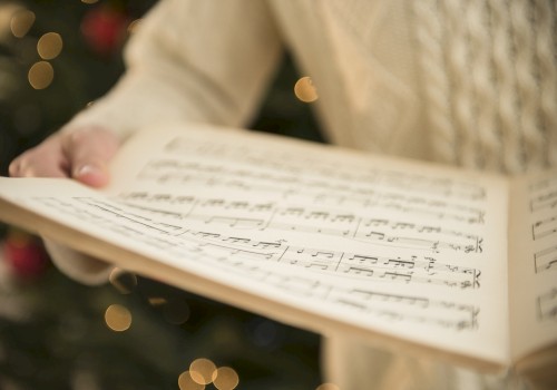 A person is holding an open book of sheet music, with out-of-focus festive lights in the background, possibly indicating a holiday season.