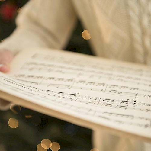 A person is holding an open book of sheet music, with out-of-focus festive lights in the background, possibly indicating a holiday season.