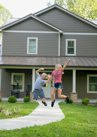 Two children are jumping and playing in front of a modern gray house with a metal roof, surrounded by greenery and trees in the background.