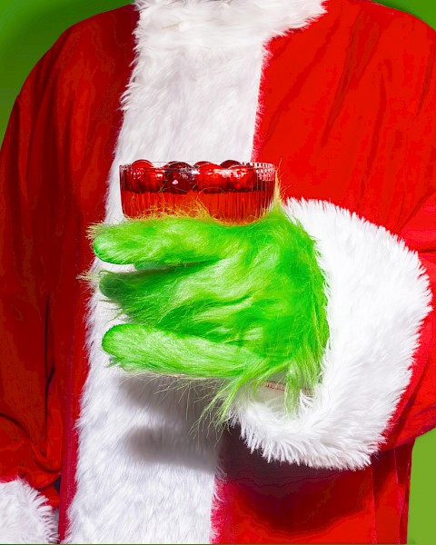 A person in a Santa suit with a green fur-covered hand holding a red cocktail against a green background.