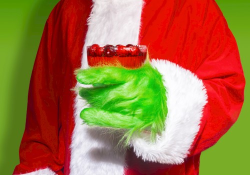 A person in a Santa suit with a green fur-covered hand holding a red cocktail against a green background.