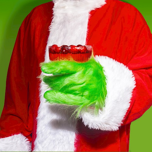 A person in a Santa suit with a green fur-covered hand holding a red cocktail against a green background.