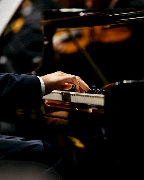 A person is playing a grand piano, with an orchestra blurred in the background. The focus is on the pianist's hands on the piano keys.