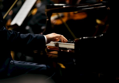 A person is playing a grand piano, with an orchestra blurred in the background. The focus is on the pianist's hands on the piano keys.