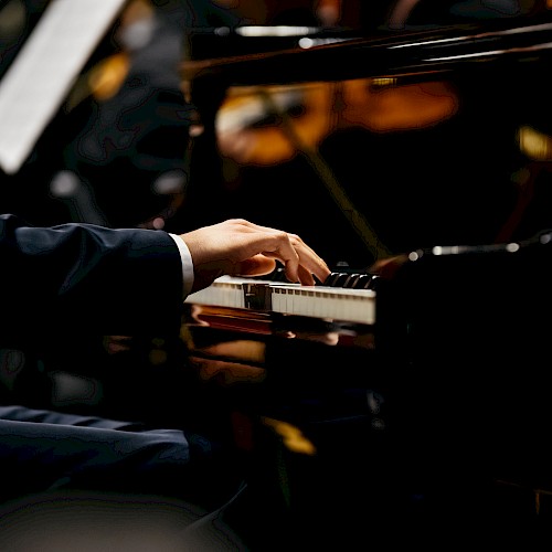 A person is playing a grand piano, with an orchestra blurred in the background. The focus is on the pianist's hands on the piano keys.