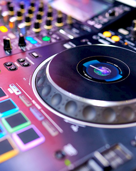 The image shows a close-up of a DJ controller with colorful pads, dials, and a large jog wheel, used for mixing music.