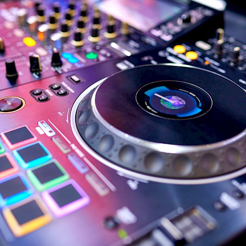 The image shows a close-up of a DJ controller with colorful pads, dials, and a large jog wheel, used for mixing music.