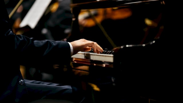 A person's hand is playing the piano, with musicians and their instruments visible in the background, suggesting a live performance or concert.
