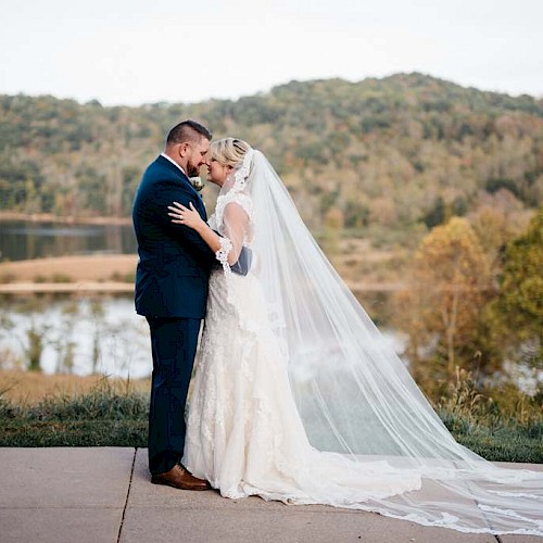 Bride and groom share an intimate moment outdoors near a lake surrounded by autumn trees.