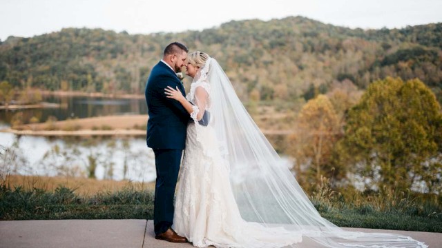 Bride and groom share an intimate moment outdoors near a lake surrounded by autumn trees.