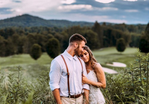 A couple stands in a lush field, embracing tenderly, with a scenic background of trees and mountains under a cloudy sky.