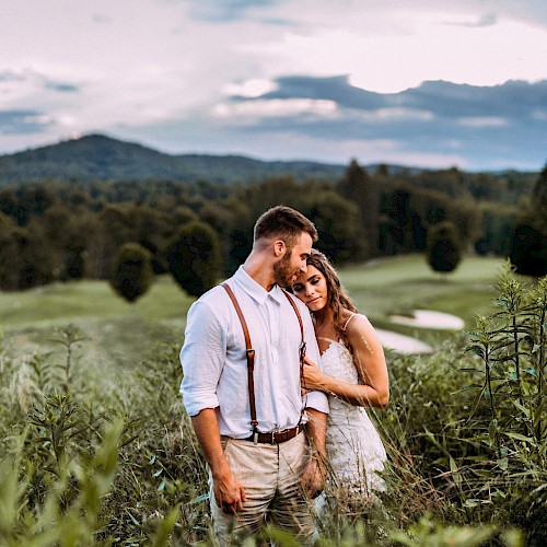 A couple stands in a lush field, embracing tenderly, with a scenic background of trees and mountains under a cloudy sky.