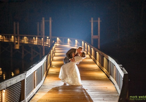 A newlywed couple shares a romantic dip on a lit wooden bridge at night, soft glow, water dark beyond, affectionate moment.