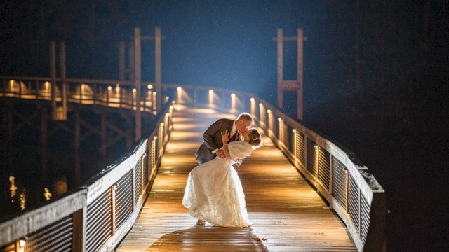 A newlywed couple shares a romantic dip on a lit wooden bridge at night, soft glow, water dark beyond, affectionate moment.
