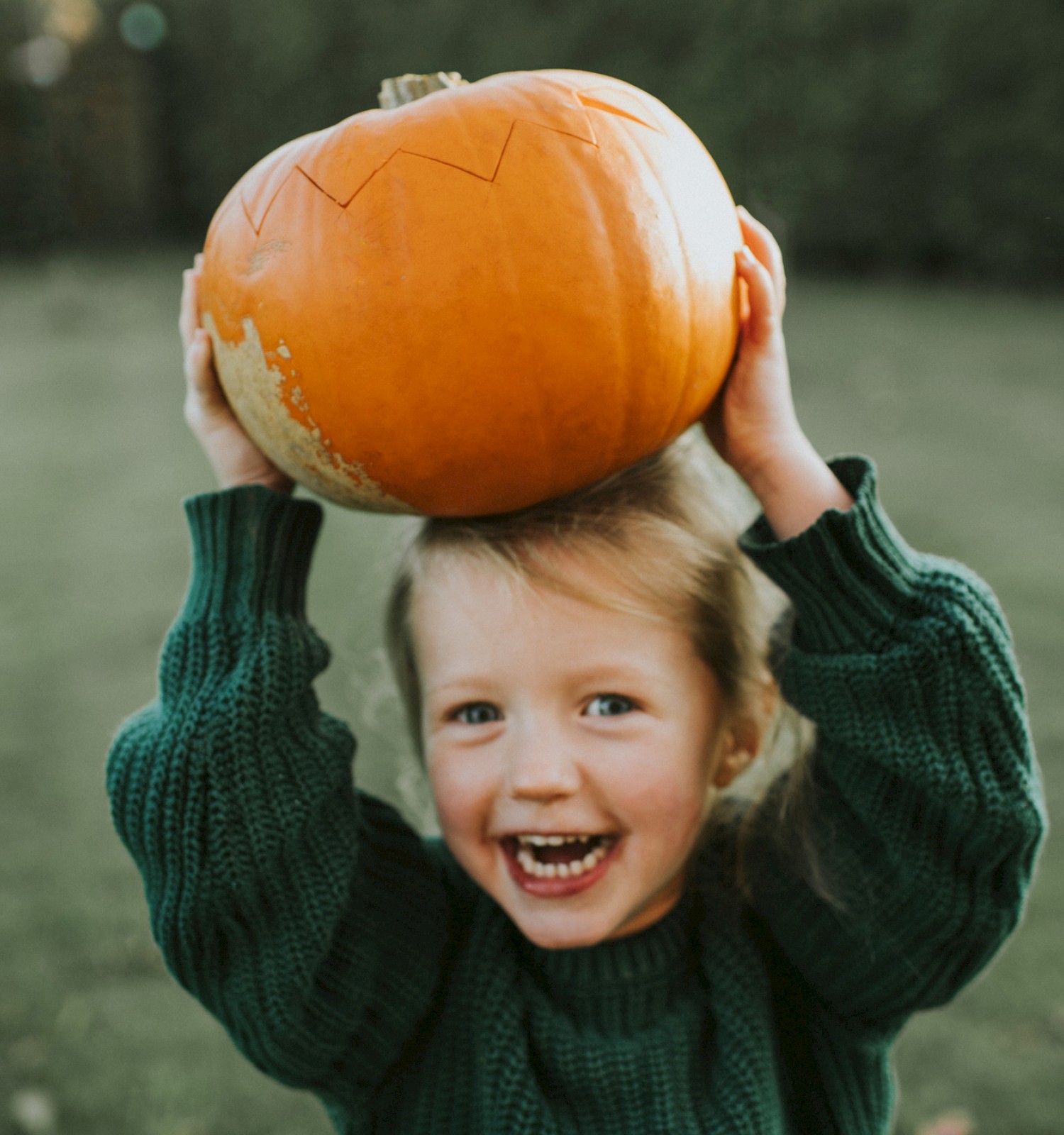 A child in a green sweater is happily holding a pumpkin above their head in a grassy area.