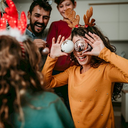 A group of people enjoying a festive moment indoors, with one person holding ornaments to their eyes, and others smiling in the background.