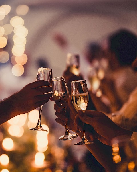 People are holding and clinking champagne glasses at a celebration, with warm, blurred lights in the background.