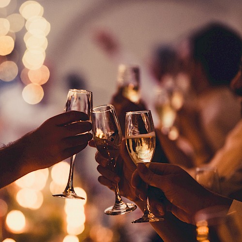 People are holding and clinking champagne glasses at a celebration, with warm, blurred lights in the background.