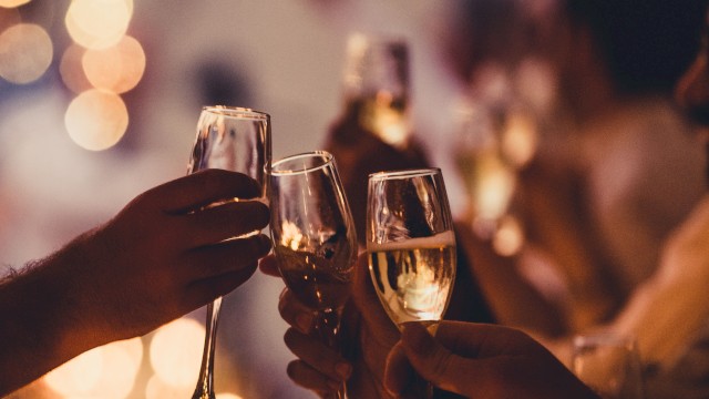 People are holding and clinking champagne glasses at a celebration, with warm, blurred lights in the background.