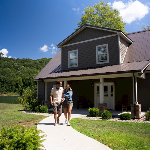 A couple walks along a curved concrete path toward a brown two-story house with a metal roof, porch, and green lawn under a sunny sky.