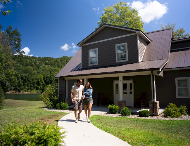 A couple walks along a curved concrete path toward a brown two-story house with a metal roof, porch, and green lawn under a sunny sky.