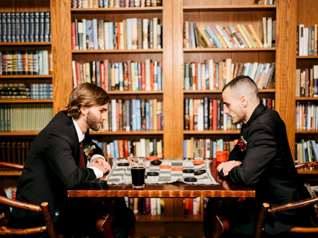 Two people in suits are playing checkers in a library, surrounded by bookshelves and with drinks on the table.