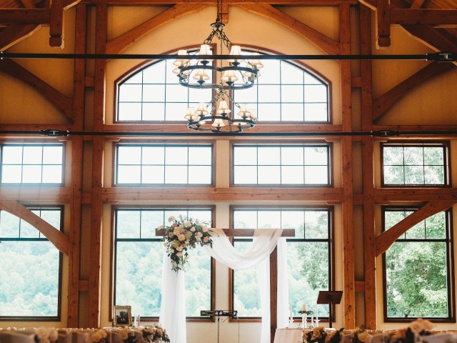A beautifully decorated indoor wedding venue with wooden beams, chandeliers, and rows of chairs, leading to a floral arch at the front.