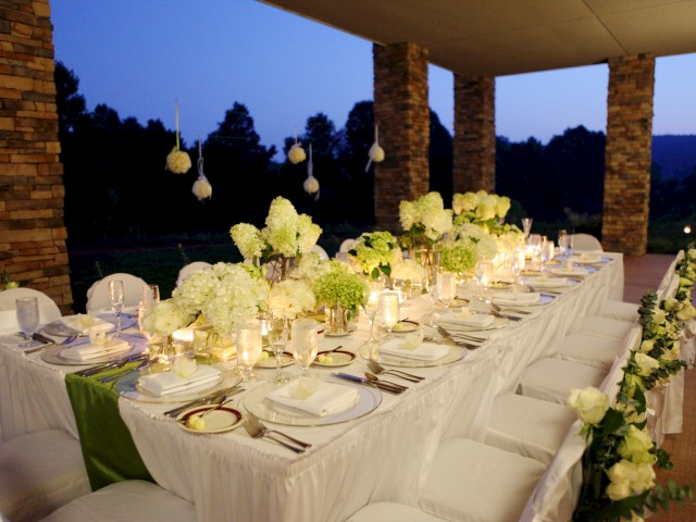 An elegantly set outdoor dinner table with white flowers, candles, and neatly arranged place settings, against a scenic dusk backdrop.