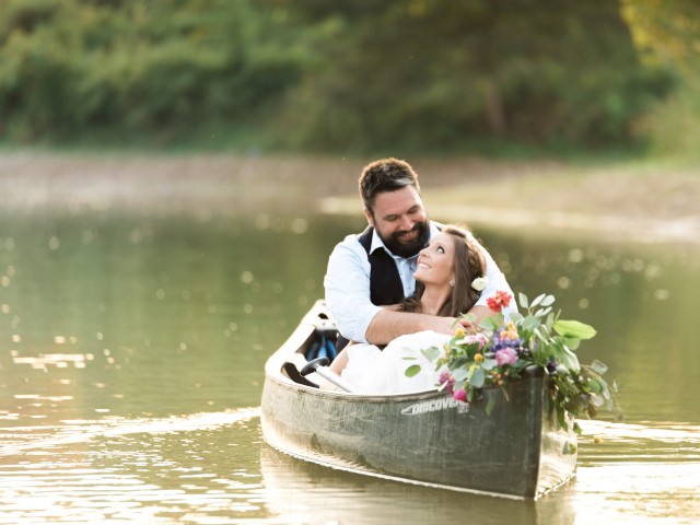 A couple is sitting in a small boat on calm water, smiling at each other. The boat is decorated with flowers, creating a romantic scene.