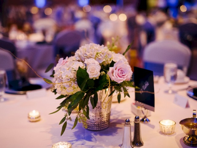 A floral centerpiece with roses and hydrangeas on a table, surrounded by candles, salt and pepper shakers, and table settings in a softly lit room.