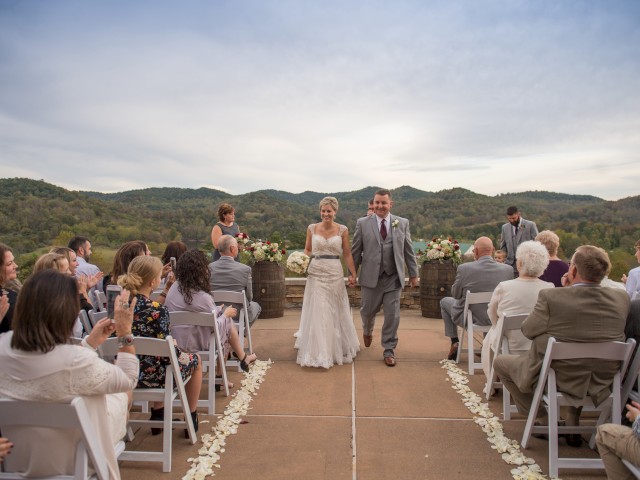 A couple walking down the aisle at an outdoor wedding ceremony, surrounded by guests and set against a scenic backdrop of hills and trees.