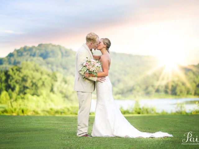 A couple in wedding attire kisses on a grassy hill with a scenic background and sunset, holding a bouquet.